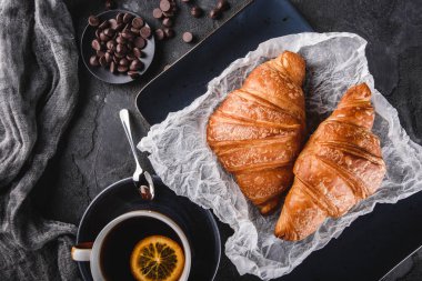 Taze Fransız çikolatalı kruvasan ile kağıt üzerinde siyah arka plan üzerinde peçete ve fincan çay ile kahvaltı. Tatlı, puf hamur işleri, üst görünüm