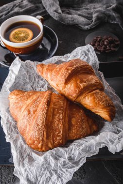Taze Fransız çikolatalı kruvasan ile kağıt üzerinde siyah arka plan üzerinde peçete ve fincan çay ile kahvaltı. Tatlı, puf hamur işleri, üst görünüm
