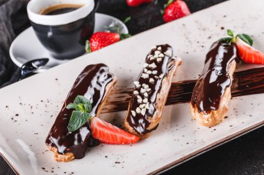 Koyu arkaplanda kremalı, çikolatalı ve çilekli ev yapımı kek eklerleri veya profiteroller bir fincan kahveyle servis edilir. Geleneksel Fransız ekler, tatlı. Üst manzara, düz uzanma