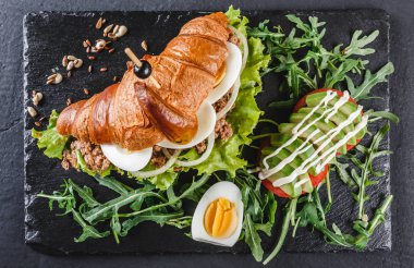 Ton balığı, yumurta, avokado, taze arugula ve siyah şist tahta üzerinde siyah taş arka plan üzerinde yeşillik ile Kruvasan sandviç. Sağlıklı gıda kavramı, Üst görünüm, düz yatıyordu