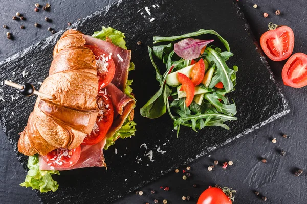 Sandwich de croissant con jamón, tomates, queso parmesano y verduras en pizarra de esquisto negro sobre fondo de piedra negra. concepto de comida saludable, vista superior, plano lay — Foto de Stock