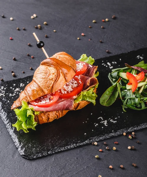 Sandwich de croissant con jamón, tomates, queso parmesano y verduras en pizarra de esquisto negro sobre fondo de piedra negra. Concepto de comida saludable — Foto de Stock