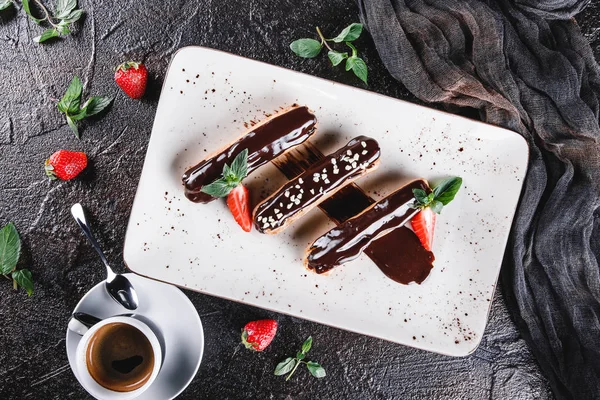 Hembakade kaka eklair eller profiteroles med vaniljsås, choklad och jordgubbar på mörk bakgrund serveras med kopp kaffe. Traditionella franska läckerheter, dessert. Ovanifrån, platt ligg. — Stockfoto