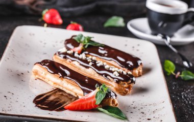 Koyu arkaplanda kremalı, çikolatalı ve çilekli ev yapımı kek eklerleri veya profiteroller bir fincan kahveyle servis edilir. Geleneksel Fransız ekler, tatlı. Yakın çekim..