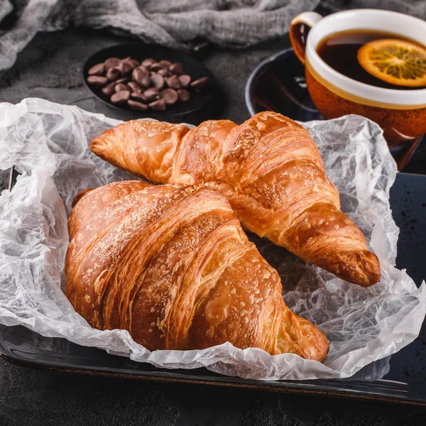 Reggeli friss francia csokis croissant a papíron több mint sötét háttér szalvétára és csésze teát. Desszert, puff péksütemény, Vértes. — Stock Fotó