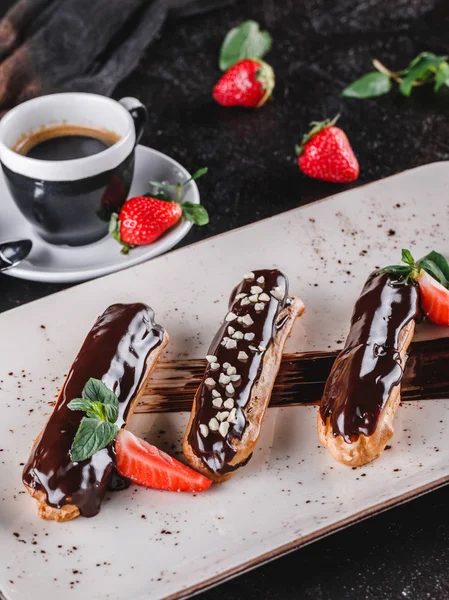 Hembakade kaka eklair eller profiteroles med vaniljsås, choklad och jordgubbar på mörk bakgrund serveras med kopp kaffe. Traditionella franska läckerheter, dessert. Närbild. — Stockfoto