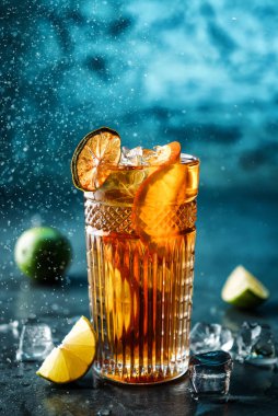 Kola, kireç, portakal ve koyu mavi arka plan üzerinde cam buz ile taze kokteyl. Stüdyoda dondurucu hareketle içki çekimi, sıvı sıçraması. Yaz soğuk içecek ve kokteyl