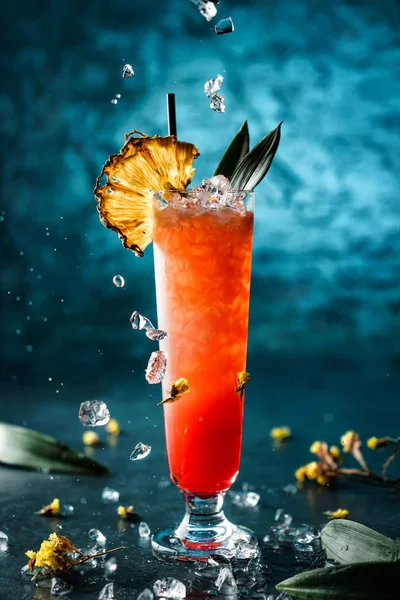 Cocktail tropical fresco com abacaxi e gelo em vidro jar sobre fundo azul escuro. Estúdio tiro de bebida em movimento de congelamento, gelo voador, flores, gotas em respingo líquido. Bebida fria de verão e coquetel — Fotografia de Stock