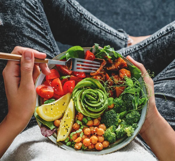 Frau in Jeans hält Buddha-Schüssel mit Salat, gebackenen Süßkartoffeln, Kichererbsen, Brokkoli, Gemüse, Avocado, Sprossen in den Händen. Gesunde vegane Ernährung, saubere Ernährung, Diät, Draufsicht — Stockfoto