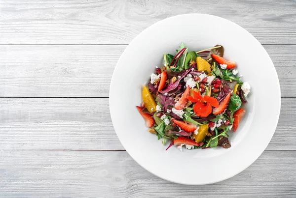 Salada fresca sã com verdes de mistura, morango, cor-de-laranja, nozes, queijo de creme e flores comestíveis na chapa no contexto de madeira leve. Comida saudável, dieta vegetariana, vista superior — Fotografia de Stock