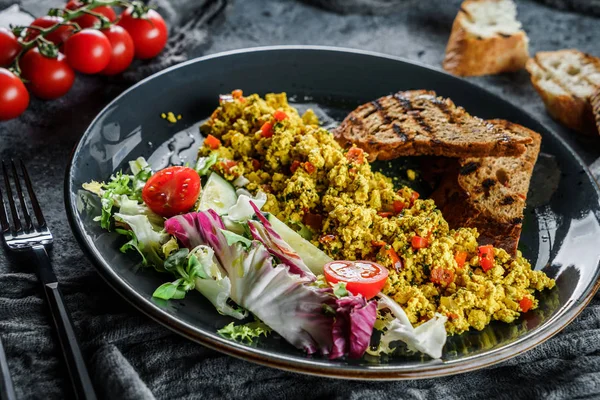 Vegan tofu scramble with vegetables, salad and toasted bread in plate over grey background. Healthy breakfast food, clean eating, vegan dieting, top view