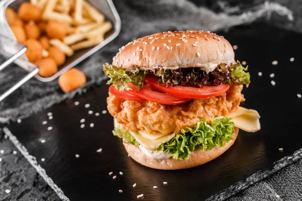 Hambúrguer de frango com queijo, tomates, alface, molho e bolas de pau frito, batatas fritas na ardósia fundo preto, close up — Fotografia de Stock