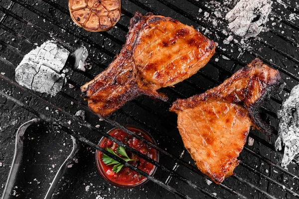 Viande grillée de steak T-bone avec ail sur les charbons sur un barbecue, fond sombre avec lumière du feu. Vue du dessus — Photo