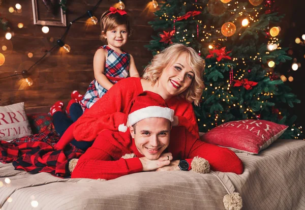 Familjen i jultomten hattar liggande på sängen. Mor, far och barn har roligt i vardagsrummet dekorerat med julgran och presentaskar. Nyår och jul semester — Stockfoto