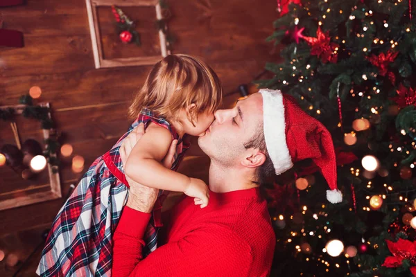 God Jul och Gott Nytt År! Far kysser sin dotter i famnen i vardagsrummet dekorerad med julgran och julklappslådor. Porträtt kärleksfull familj på nära håll, semester — Stockfoto