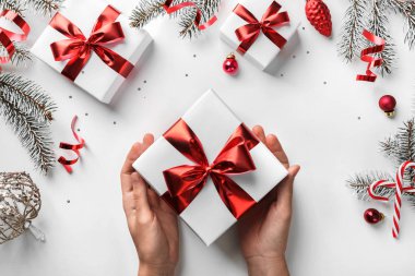 Beyaz arka planda Noel hediyesi kutusu tutan kadın eller, köknar dalları, kırmızı süslemeler, ışıltılar ve konfetiler. Noel ve yeni yıl tebrik kartı, kış tatili. Düz yatış, üst görünüm