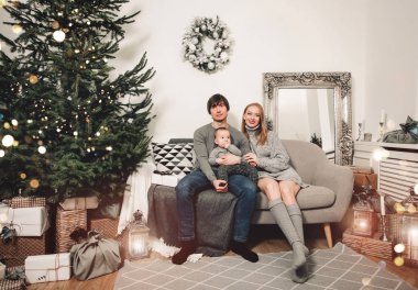 Noel ailesi ve Noel ağacının yanında gülümseyen bir bebek. Noel ağacı ve hediye kutularıyla dekore edilmiş oturma odası. Işık rahat bir ortam sağlıyor. Yeni yıl tatili