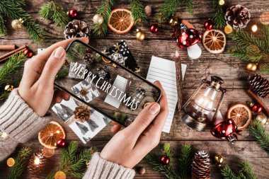 Cep telefonu tutan kadın eller. Süslü Noel hediye kutuları, dallar, tatil arka planında çam kozalakları. Xmas ve Mutlu Yıllar teması. Düz yatış, üst görünüm