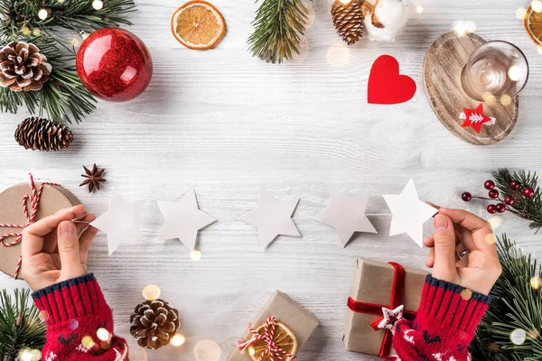 Main féminine tenant une guirlande avec des étoiles sur fond de Noël avec des branches d'arbre, des cônes de pin, des cadeaux. Noël et bonne année vacances, bokeh, lumière. Couché plat, vue du dessus — Photo
