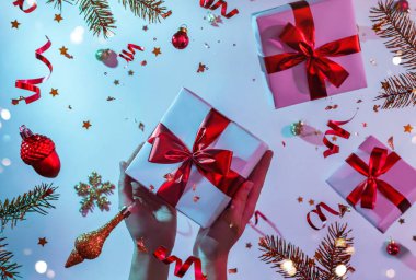 Bayanların elinde renkli parlak neon rengi parlak mavi ve mor arka planda uçan Noel süslemeleri, köknar dalları ve konfetileri olan hediye kutusu var. Noel ve Yeni Yıl tatili, Bokeh, ışık. Seçici odak