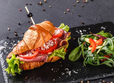 Prosciutto, domates, peynirli parmesan peynirli kruvasan sandviçi ve siyah taş arka planda siyah şist tahtası. Sağlıklı gıda konsepti