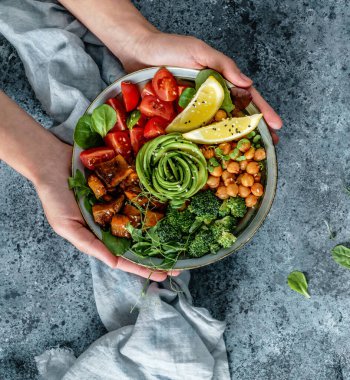 Ellerinde sağlıklı bir süper kase ya da salatalı Buda kasesi fırında patates, nohut, brokoli, humus, avokado, açık mavi arka planda lahanalar. Sağlıklı vejetaryen yemekleri, temiz yemek, üst manzara. 