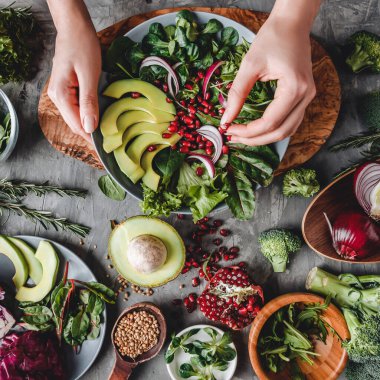 Kadın avokado, yeşillik, roka ve gri arka planda ıspanak ile sağlıklı taze salata pişiriyor. Sağlıklı vejetaryen yemekleri, temiz yemek, diyet, üst görünüm, ton.