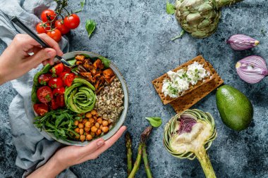 Ellerinde sağlıklı bir süper kase salata, tatlı patates, nohut, kinoa, domates, roka ve açık mavi arka planda avokado olan Buda kasesi var. Sağlıklı vejetaryen yemekleri, temiz yemek, üst manzara.