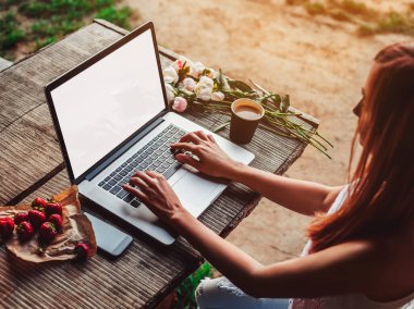 Kahve fincanı, çilek, şakayık çiçeği ve akıllı telefonlu kaba ahşap masada dizüstü bilgisayar kullanan ve daktilo kullanan genç bir kadın. Açık hava parkında çalışan serbest çalışan biri. Tonlama