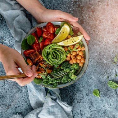 Ellerinde sağlıklı bir süper kase ya da salatalı Buda kasesi fırında patates, nohut, brokoli, humus, avokado, açık mavi arka planda lahanalar. Sağlıklı vejetaryen yemekleri, temiz yemek, üst manzara. 