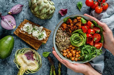 Ellerinde sağlıklı bir süper kase salata, tatlı patates, nohut, kinoa, domates, roka ve açık mavi arka planda avokado olan Buda kasesi var. Sağlıklı vejetaryen yemekleri, temiz yemek, üst manzara.
