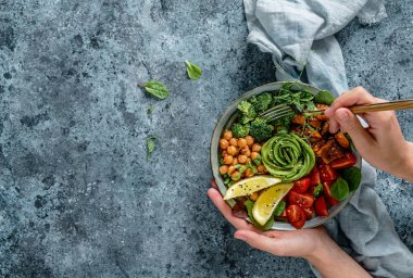 Ellerinde sağlıklı bir süper kase ya da salatalı Buda kasesi fırında patates, nohut, brokoli, humus, avokado, açık mavi arka planda lahanalar. Sağlıklı vejetaryen yemekleri, temiz yemek, üst manzara. 