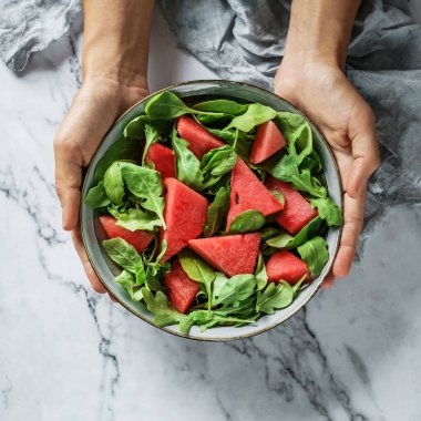 Elleri sağlıklı taze karpuz salatası, roka, ıspanak ve açık mermer arka planda yeşillikler tutuyor. Sağlıklı yemek, temiz yemek, Buda kasesi salatası, üst manzara.