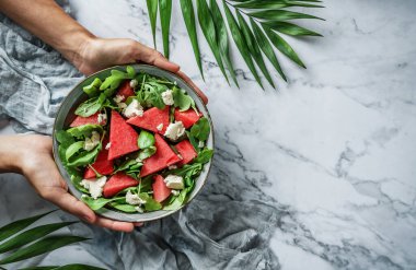 Elleri sağlıklı taze karpuz salatası, roka, ıspanak ve açık mermer arka planda tropikal yapraklı yeşillikler tutuyor. Sağlıklı yemek, temiz yemek, Buda kasesi salatası, üst manzara.