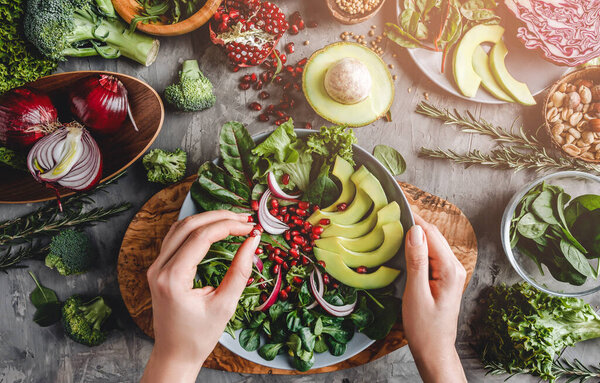 Woman cooking healthy fresh salad with avocado, greens, arugula, spinach in plate over grey background. Healthy vegan food, clean eating, dieting, top view