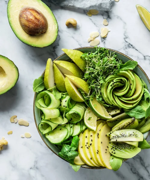 Fresh summer salad with avocado, kiwi, apple, cucumber, pear, micro greens, lime and sesame with smoothie on light marble background. Healthy food, clean eating, Buddha bowl salad, top view