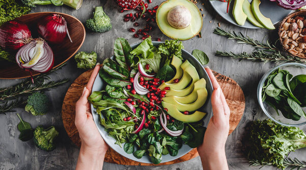 Woman cooking healthy fresh salad with avocado, greens, arugula, spinach in plate over grey background. Healthy vegan food, clean eating, dieting, top view, toning