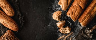 Taze ekmek baget ve çörekler siyah tahta üzerinde buğday dikenli, çuval bezli. Hamur işleri ve fırın, üst manzara, geniş kompozisyon.