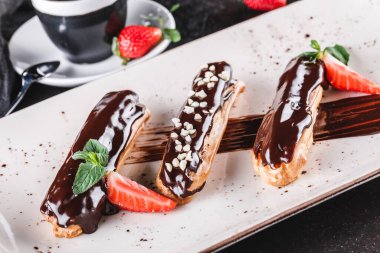 Koyu arkaplanda kremalı, çikolatalı ve çilekli ev yapımı kek eklerleri veya profiteroller bir fincan kahveyle servis edilir. Geleneksel Fransız ekler, tatlı. Yaklaş, makro.