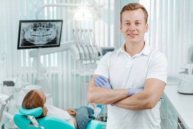 Doktor dişçi gülümsüyor ve elleri arkaplanda dişçi kliniğinde tıbbi ekipman, diş röntgeni ve hasta ile çapraz duruyor. Gülümse sağlıklı diş konsepti