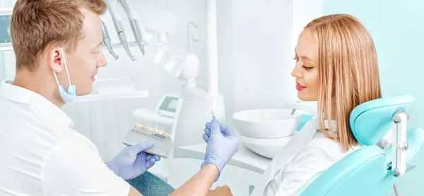 Doctor dentist chooses the whitening color for teeth the young attractive patient in stomotology clinic. Hands dentist with teeth color palette next by face. Smile healthy teeth concept