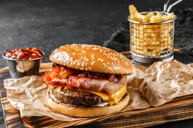 Et pirzolası, bir dilim peynir, ahşap tahtada salam ve baharatlı sos ve siyah arka planda patates kızartması ile lezzetli bir burger. Fast food öğle yemeği, kapat.