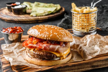 Et pirzolası, bir dilim peynir, ahşap tahtada salam ve baharatlı sos ve siyah arka planda patates kızartması ile lezzetli bir burger. Fast food öğle yemeği, kapat.