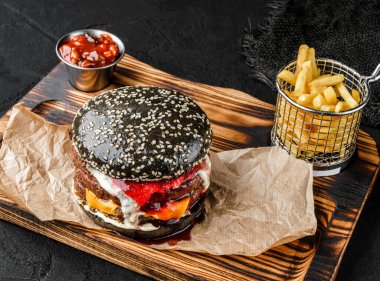 İki köfteli duble siyah burger, bir dilim peynir, mayonez ve böğürtlen sosu tahtada, siyah arka planda patates kızartması. Fast food öğle yemeği, üst manzara.