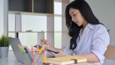 Ofiste çalışan Asyalı genç kadın elindeki bilgisayarlara ve ofis malzemelerine baktı..