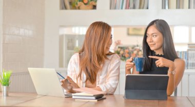 İki genç kız öğrenci gülümseyerek konuşuyor ve rapor hazırlarken kahve içiyorlar..