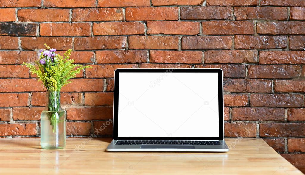 Mockup laptop blank screen with flowers in a glass jar placed on a wooden table. Red brick wall background.