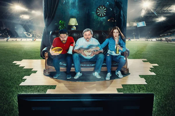 Soccer football fans sitting on the sofa and watching TV in the middle of a football field.
