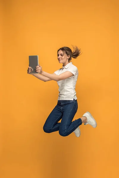 Immagine di giovane donna su sfondo arancione utilizzando computer portatile o gadget tablet durante il salto . — Foto Stock