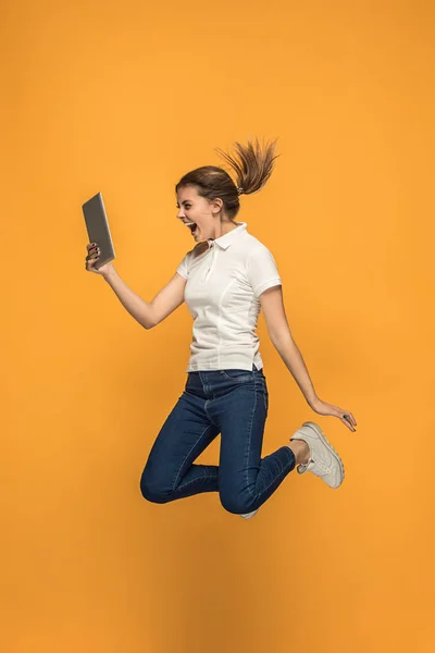 Immagine di giovane donna su sfondo arancione utilizzando computer portatile o gadget tablet durante il salto . — Foto Stock